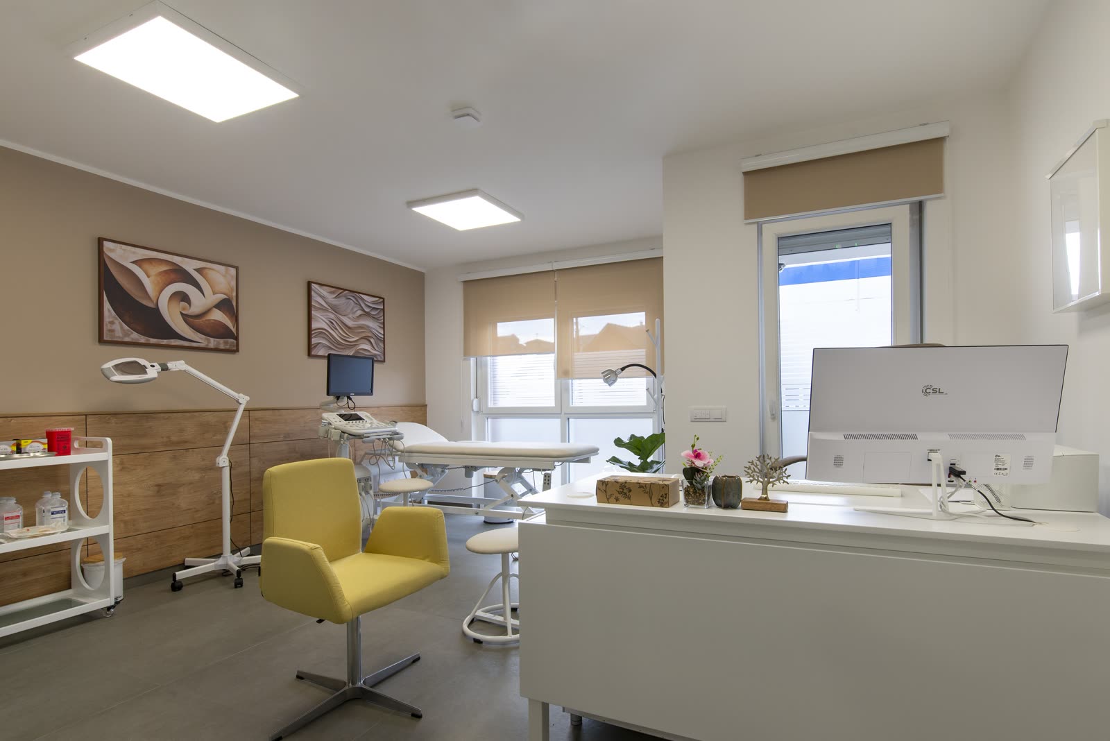 Sala de procedimentos clara, com poltrona amarela, maca, luminária e uma mesa branca